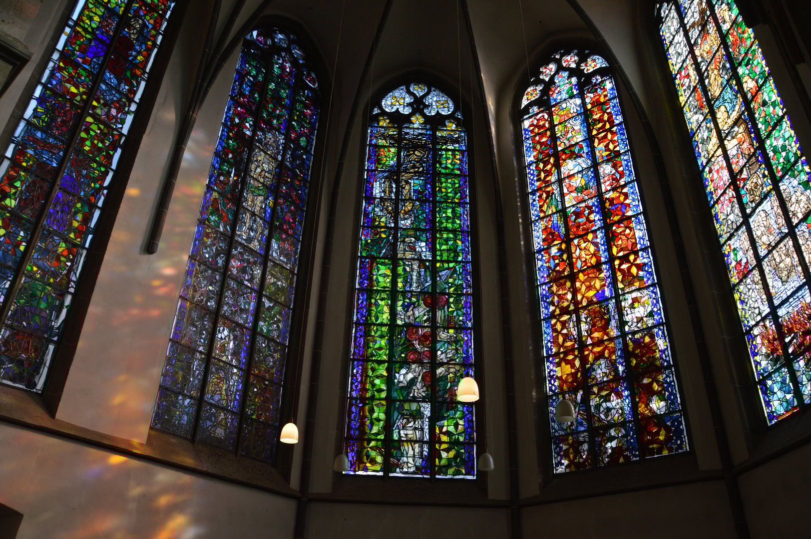 Stained glass windows in a church, featuring colorful abstract patterns with arched tops and light casting patterns on walls.