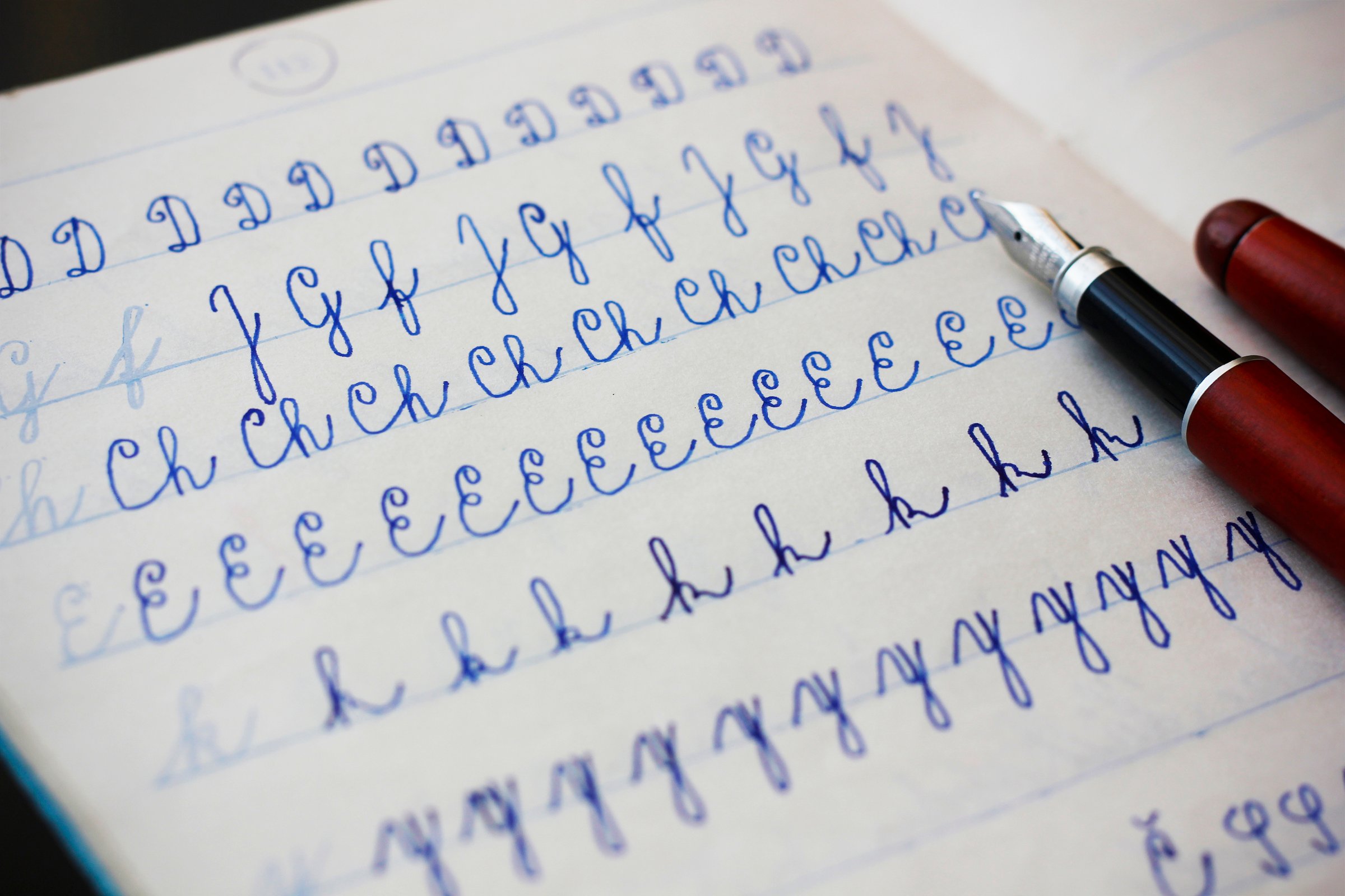 Close-up of uppercase and lowercase letters written by a child in cursive with a fountain pen with blue ink in a lined notebook, a burgundy fountain pen in the background. Concept for school education and handwritten documents.