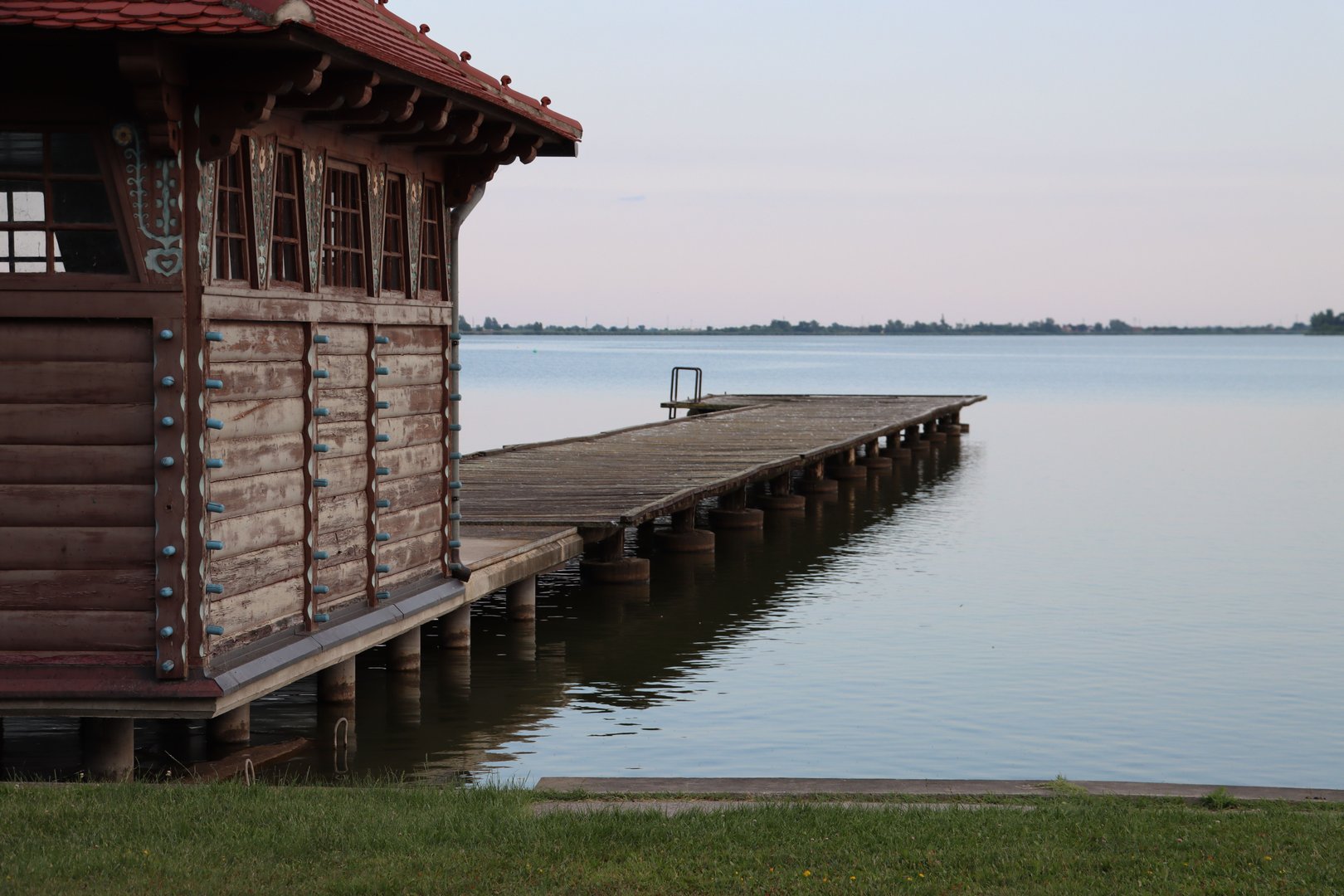 A serene lakeside view featuring a rustic wooden cabin and pier extending over calm waters under a pastel sky.