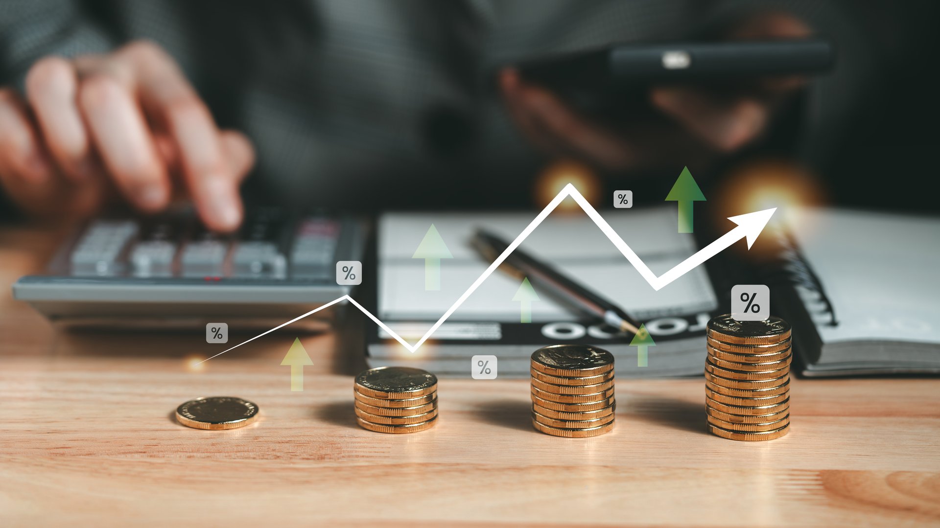 Stacks of coins and an upward graph overlay highlight financial growth, as a hand works on a calculator and notebook to plan investments and savings