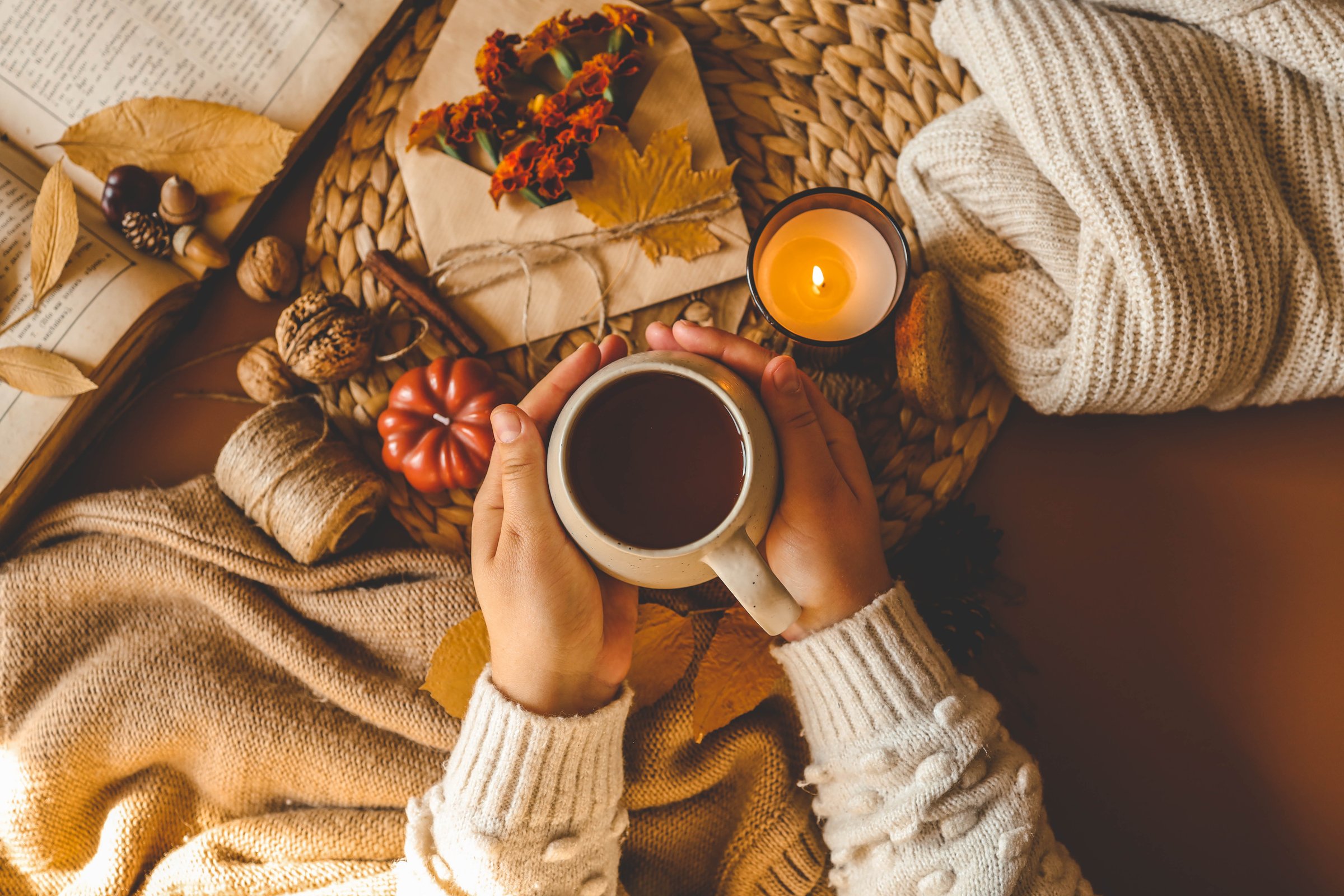 Cozy autumn flat lay featuring a warm drink held in hands, surrounded by fall elements such as a candle, pumpkin, twine, dried leaves, and a soft knitted sweater, evoking comfort and seasonal warmth.