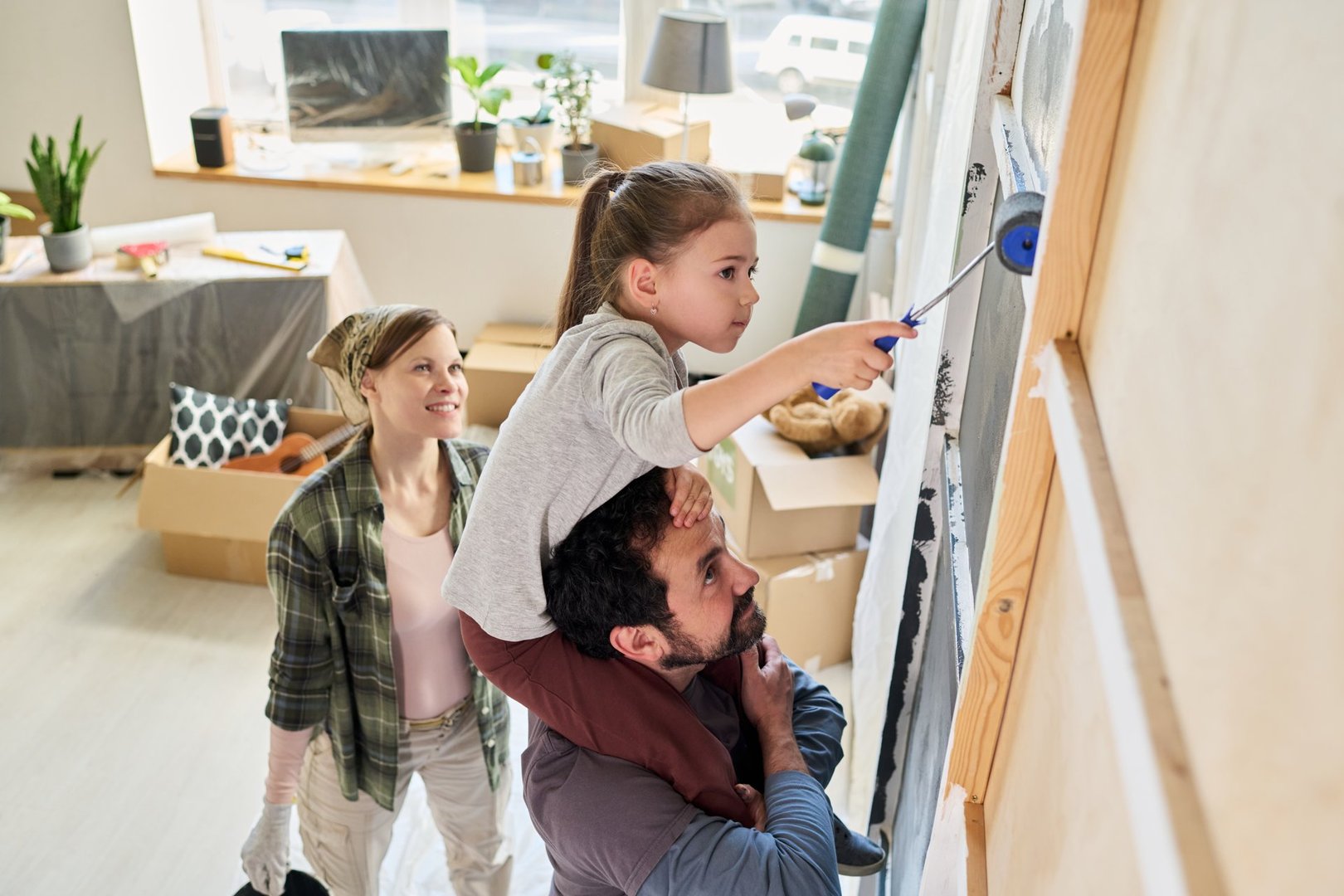 Adorable girl sitting on shoulders of young bearded man and painting wall of living room with paintroller while helping parents