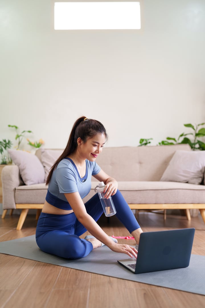 Happy Asian adult woman in sportswear on yoga mat using laptop at home. Wellness lifestyle and healthy routine activity.