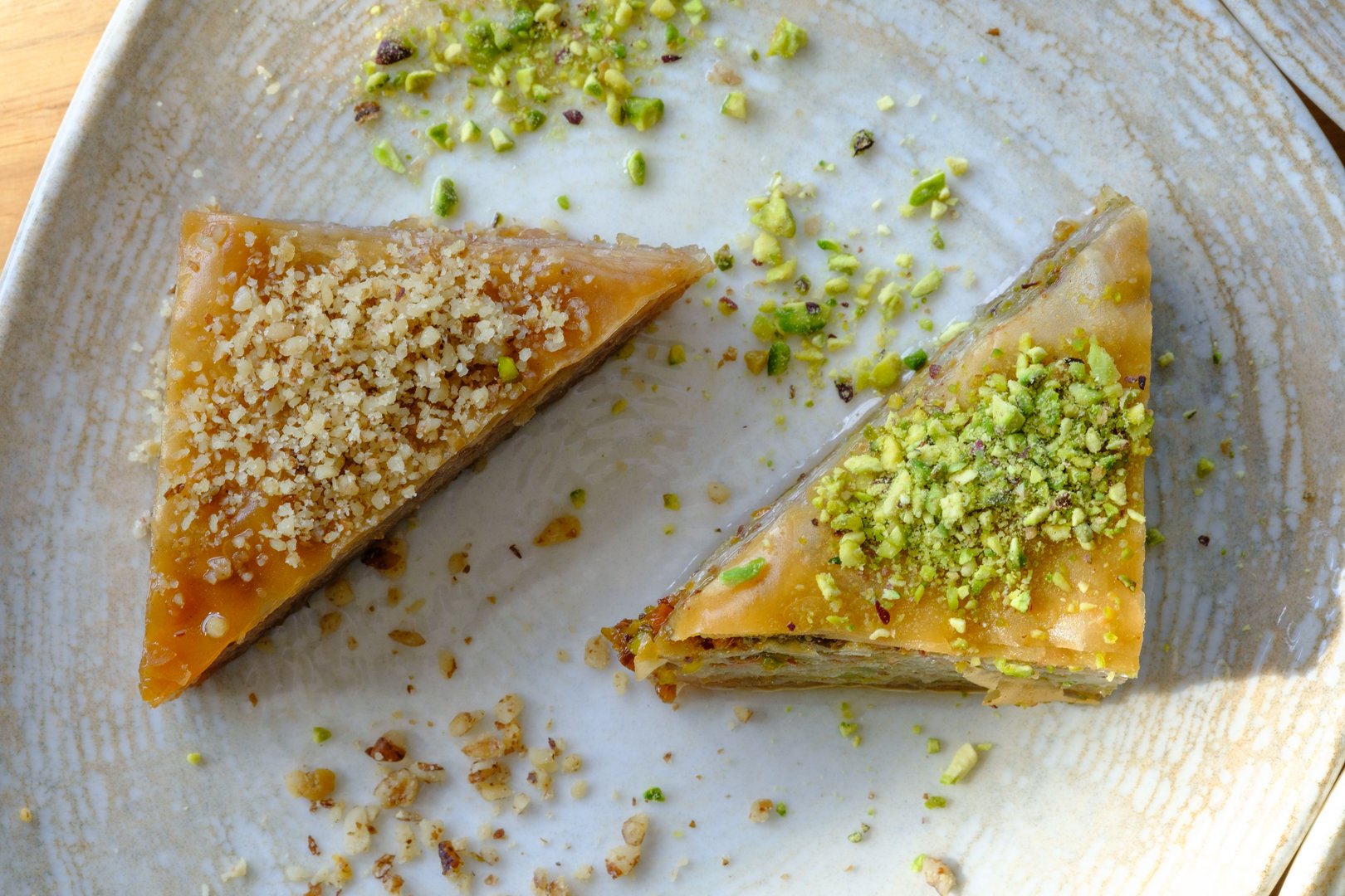 A close-up overhead shot shows two triangular pieces of baklava. One is sprinkled with chopped walnuts, the other with pistachios.  Both rest on a light-colored plate.