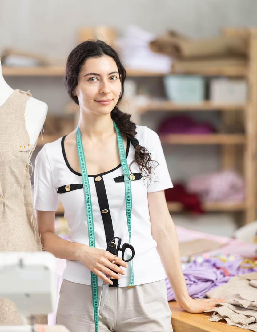 Woman dressmaker stands in a sewing workshop against the background of shelves with fabrics and sewing accessories. Seamstress smiles and invites customers to place orders