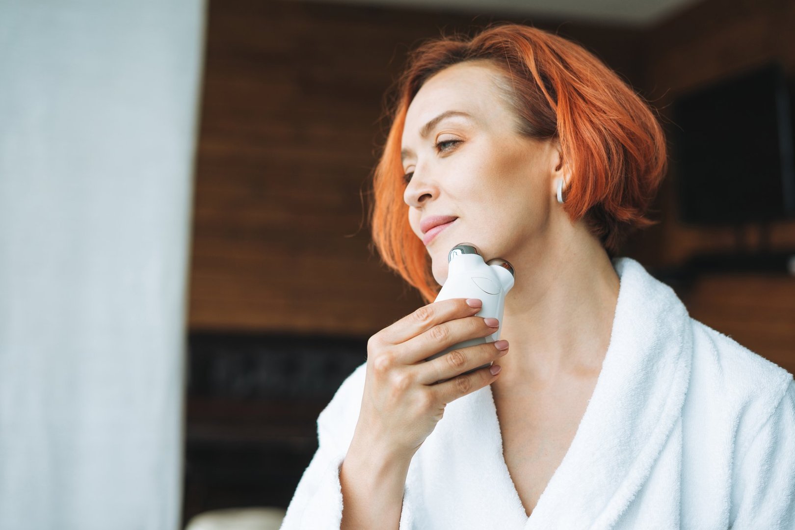 Beauty portrait of smiling woman 35 year in white bathrobe with clean fresh face and hands with red hair doing fasial massage with microcurrent facial massager at the bath room, treat yourself, home body care