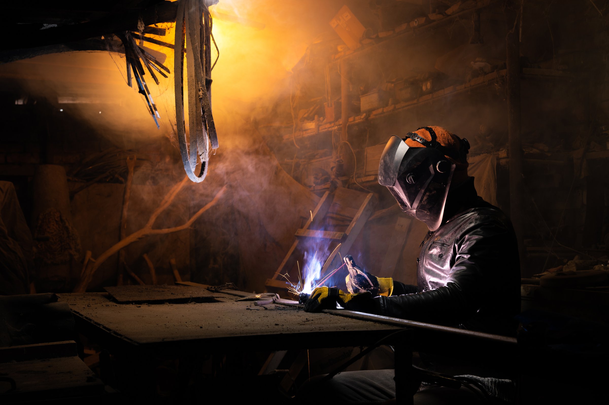 A welder in a protective mask for metal welding and protective gloves in a leather jacket performs welding work in a home garage. Sparks, hot flame from welding