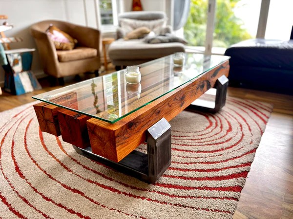 A modern coffee table with a glass top and wooden frame, set on a circular red and white patterned rug in a living room.