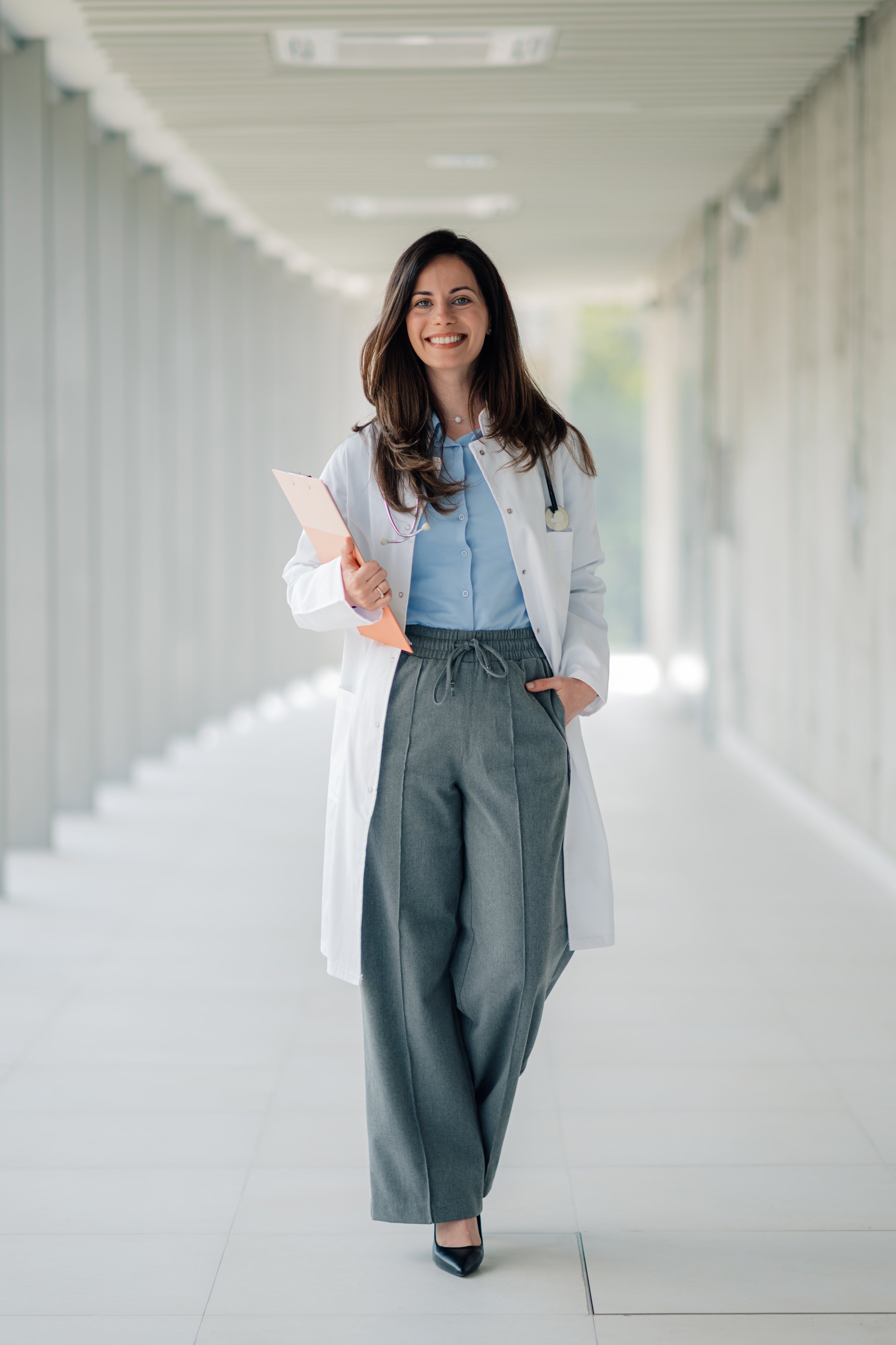 Female doctor walking confidently down a hospital corridor, holding a clipboard and smiling warmly at the camera with professionalism