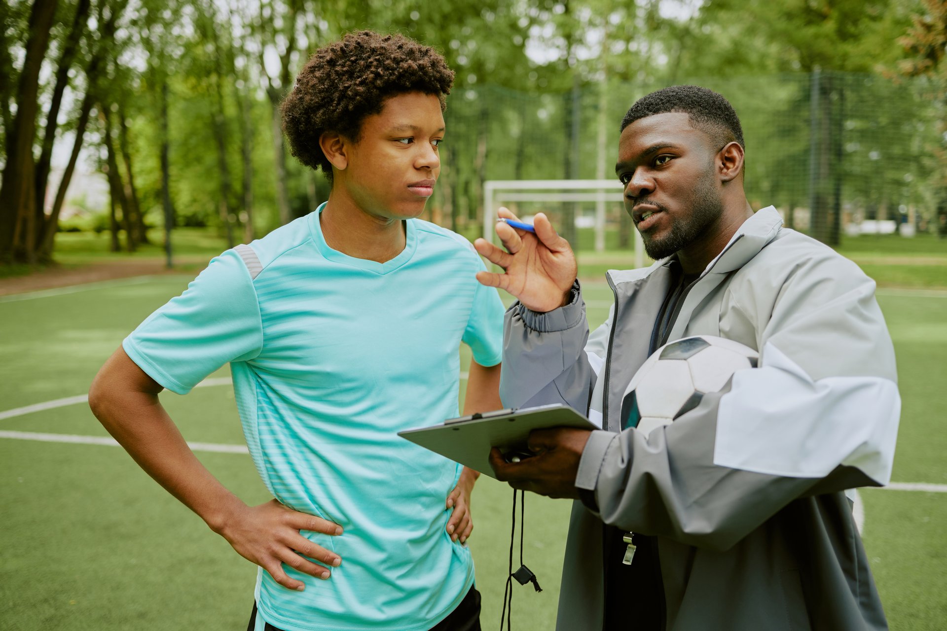 Teenager listening to Black male coach explaining strategy while holding soccer ball and clipboard on outdoor sports field