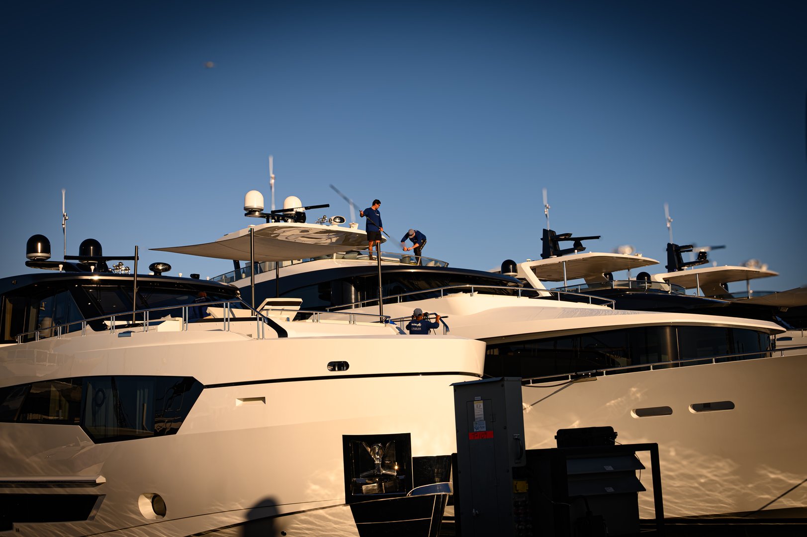 A row of gleaming luxury yachts lines the marina at the Fort Lauderdale Boat Show, with crew members on upper decks under a bright blue sky. The image conveys wealth, leisure, and nautical culture, highlighting sleek hulls, glossy finishes, and the energetic atmosphere of a premier maritime event.
