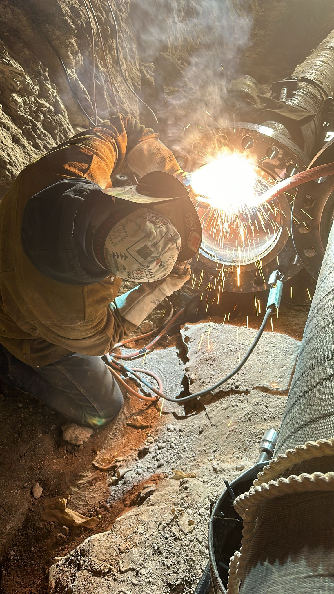 Welder in protective gear works on a pipeline joint underground, with sparks flying in dimly lit, rocky surroundings.