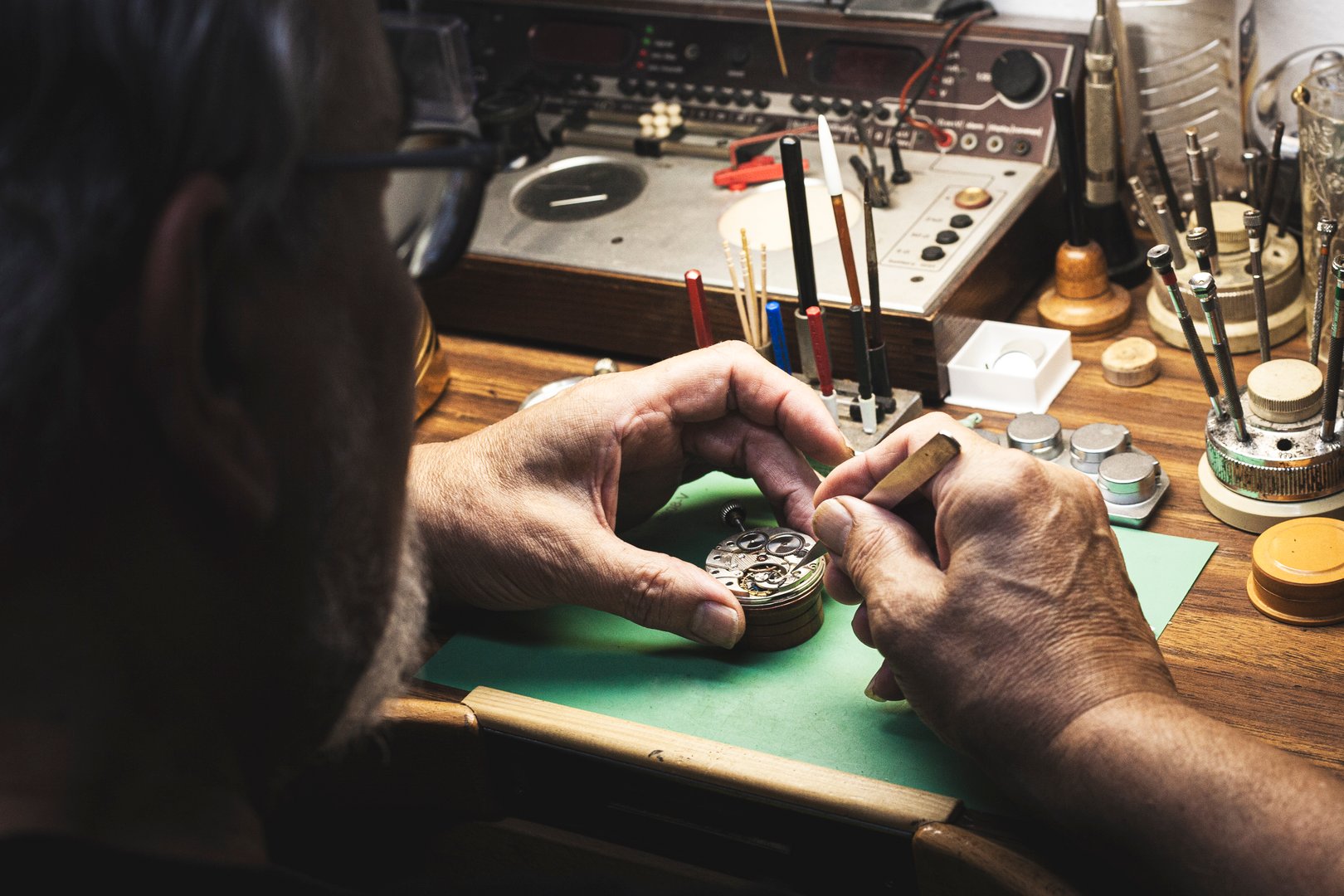 Shot from behind a watchmaker at his workbench full of tools and his expert hands are seen repairing a watch engine with a pair of tweezers.