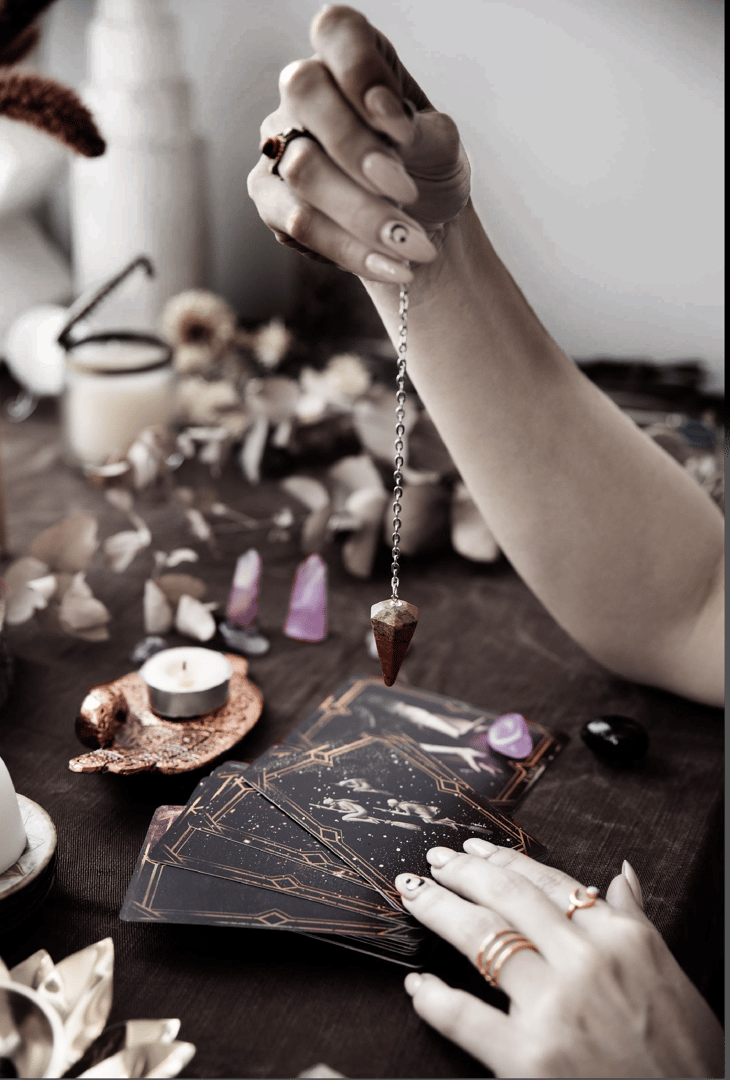 Person using a pendulum over tarot cards on a wooden table with crystals, candles, and petals around.