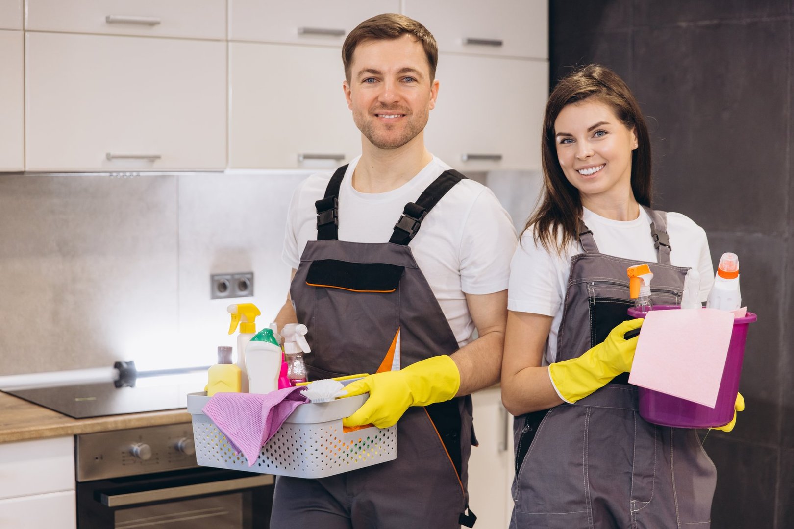 Professional cleaners holding basket and bucket with cleaning products smiling in kitchen