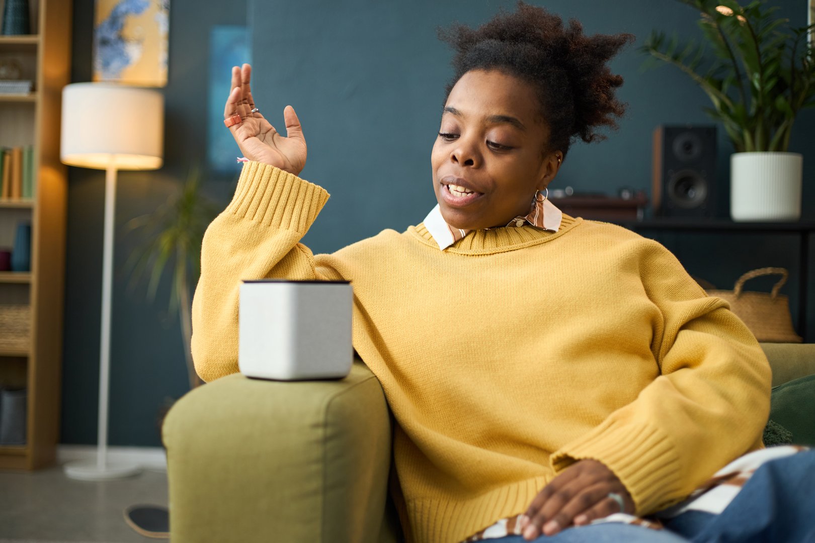 Young African American woman interacting with smart speaker at home, engaging in conversation. Comfortable and relaxed setting with modern decor and household items