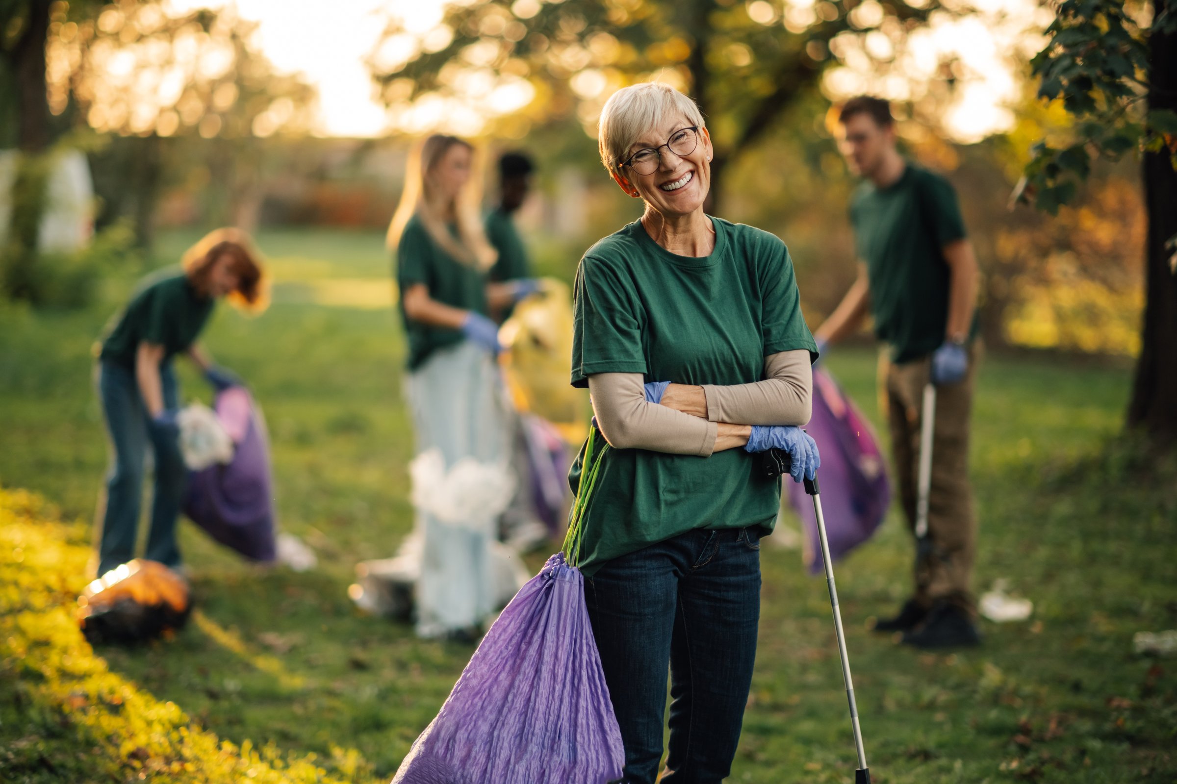 Senior woman is smiling while posing with her arms crossed during a volunteering event in a park. Her fellow volunteers are working blurred in the background