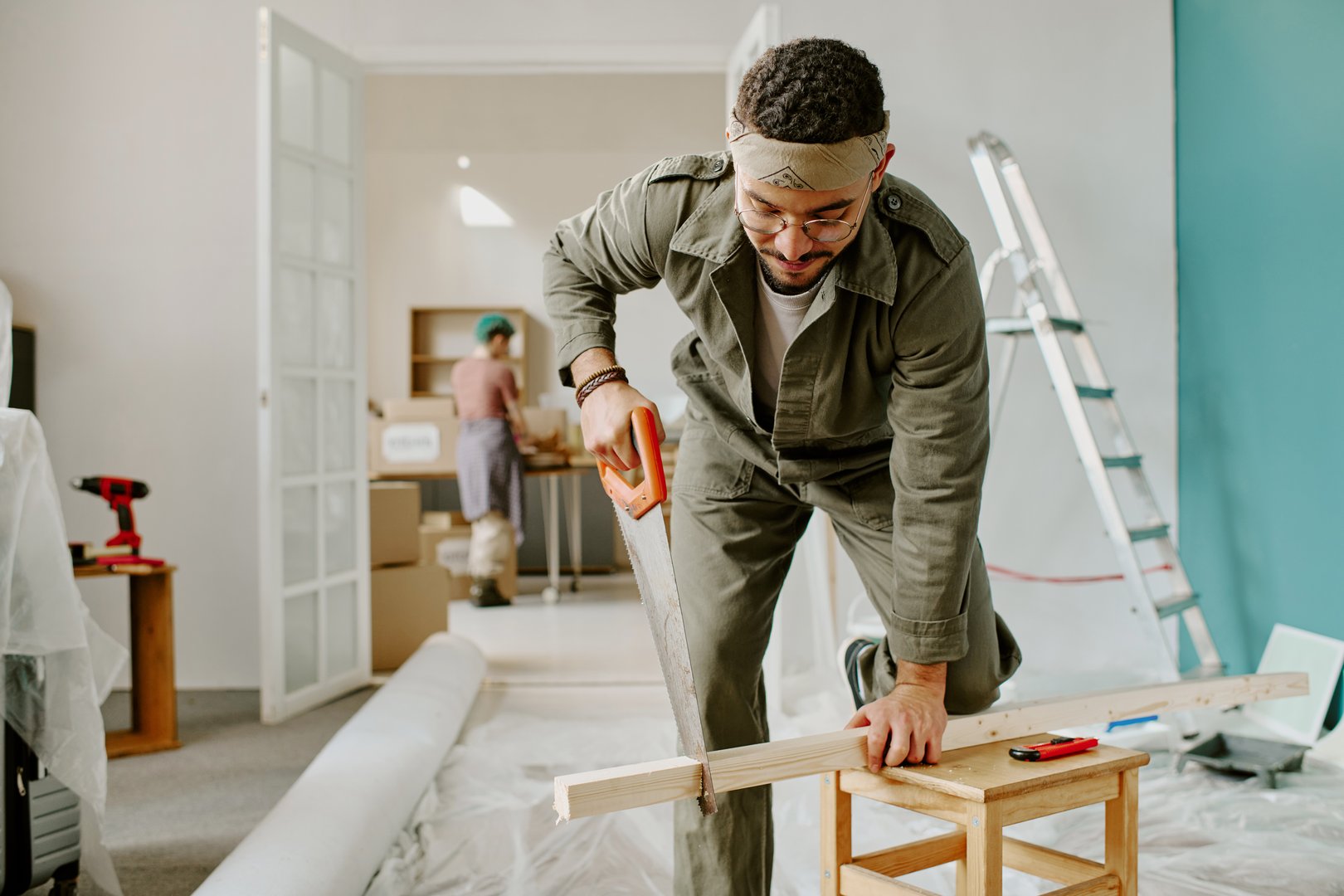 Young man sawing wooden plank while kneeling on chair, young Caucasian woman unpacking boxes in background
