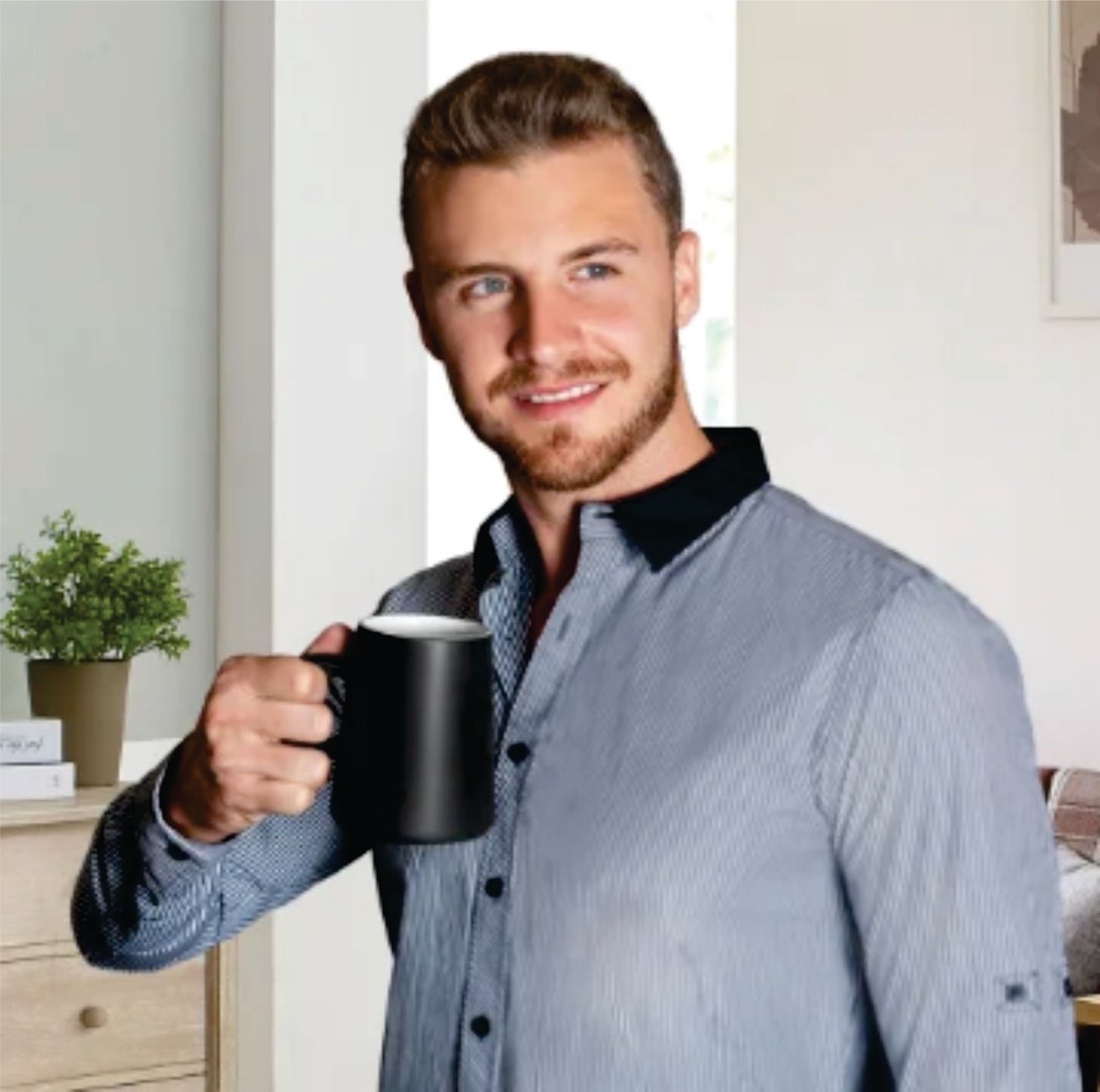 Man with a beard smiling while holding a black mug, standing in a room with light walls and a green plant in the background.