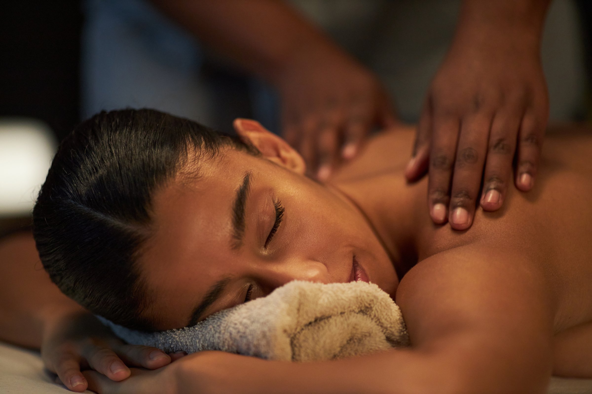 Woman lying face down, eyes closed, receiving a massage on her shoulders, towel under her head providing comfort and spa environment creating peaceful atmosphere