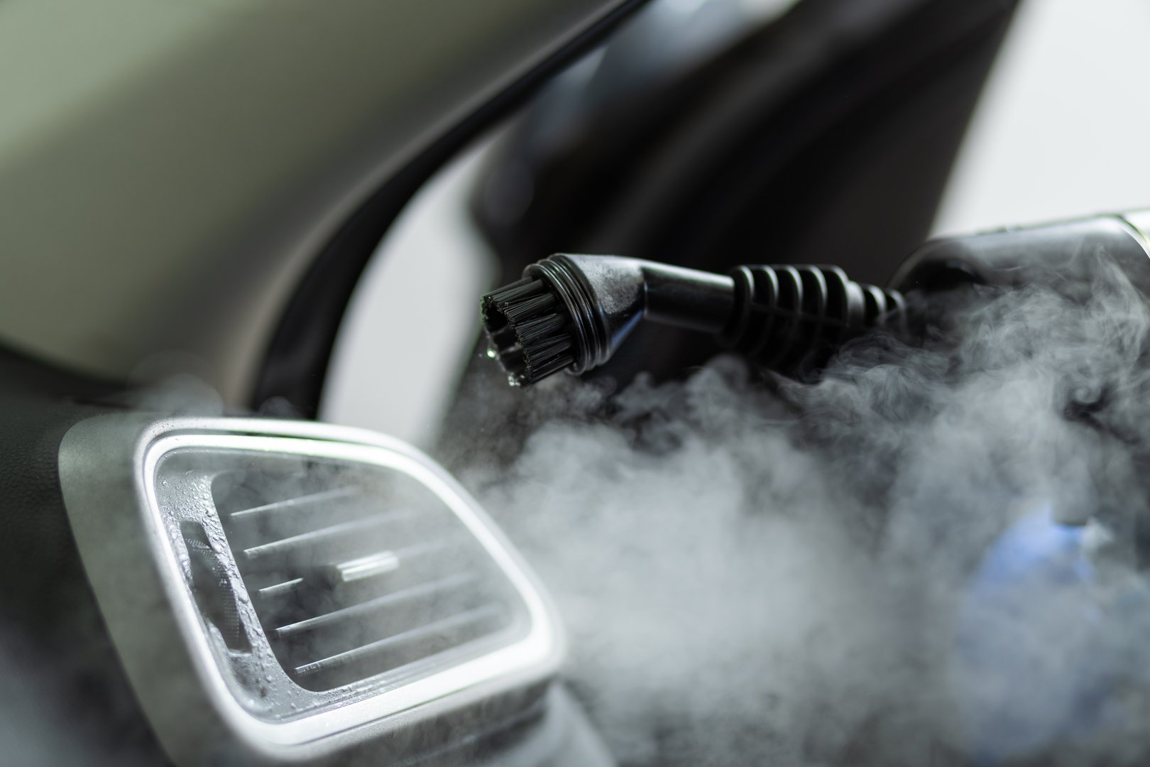 A steam cleaner nozzle is pointed at the air vent of a car. The steam is visible as it comes out of the nozzle and fills the car interior.