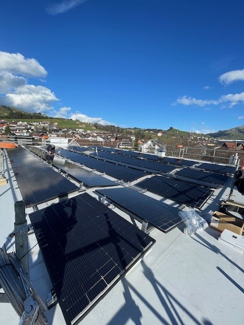 Rows of solar panels on a rooftop with a clear blue sky and a hilly landscape in the background.