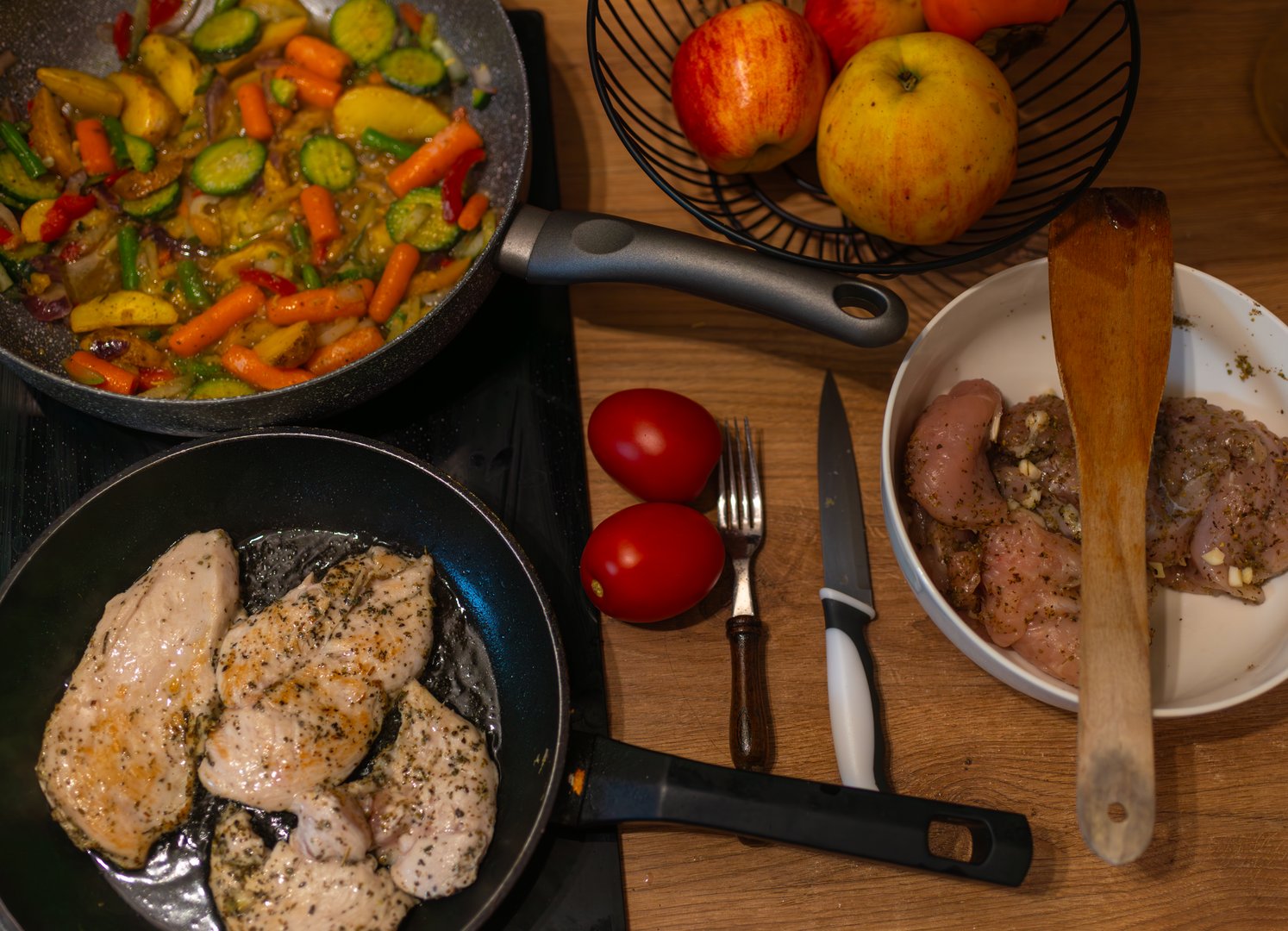 Chicken fillets sizzle in one pan while colorful vegetables cook in another. Fresh tomatoes, spices, knives and utensils complete a home-cooking scene, perfect for culinary, recipe, and food preparation themes.