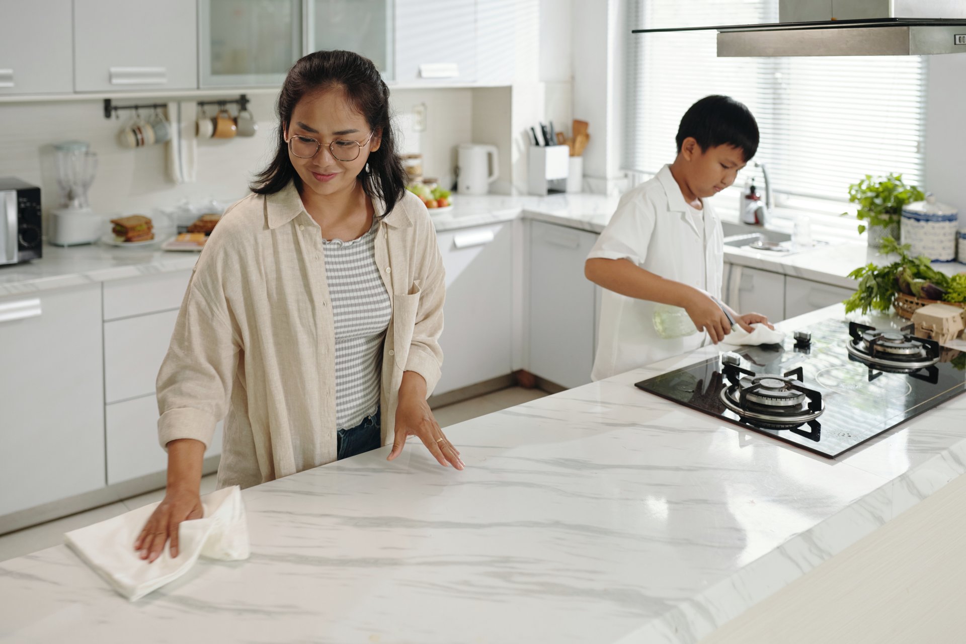 Woman cleaning marble countertop while boy cooking on stove in a bright and airy kitchen featuring modern appliances and ample counter space