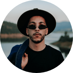 Man wearing a black hat and sunglasses with a calm expression, standing outdoors with a lake and hills in the background.