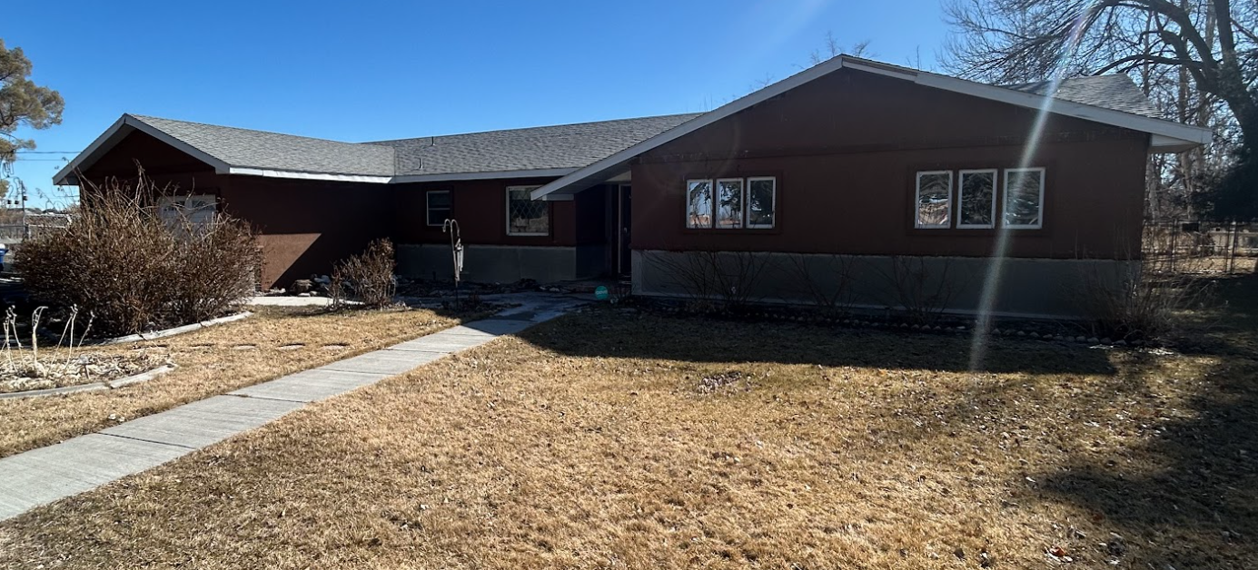 A single-story brown house with a gray roof, front lawn, and a concrete walkway under a clear blue sky.
