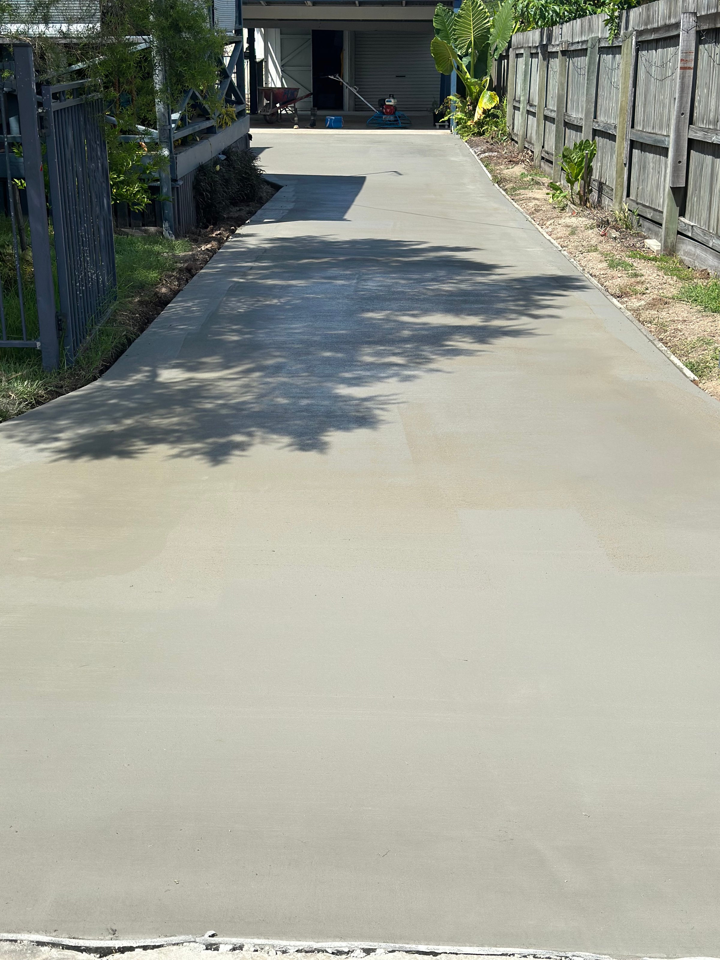 Freshly poured concrete driveway with smooth surface, flanked by a wooden fence and greenery, leading to a distant garage.