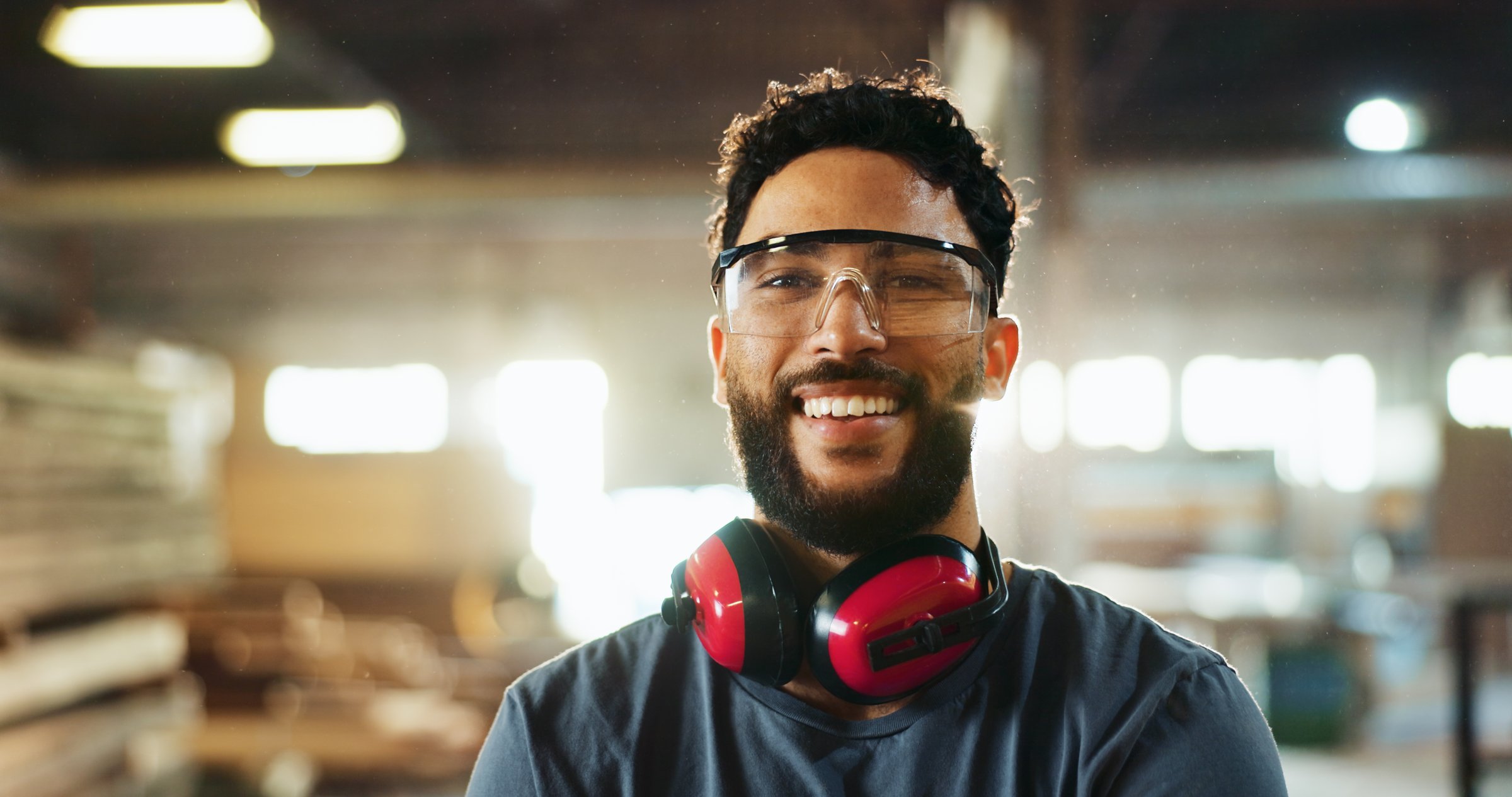 Carpenter, man or portrait with goggles in workshop for woodwork, furniture production or work safety. Manufacturing industry, headphones and happy worker in warehouse for carpentry or small business