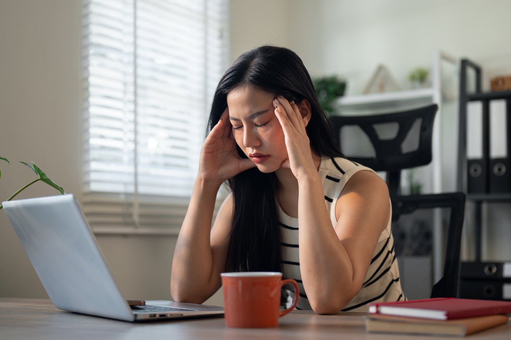 A frustrated, tired Asian businesswoman sits in front of a computer, massaging her temples, struggling with a severe headache while working, exhausted from a long day at the office.