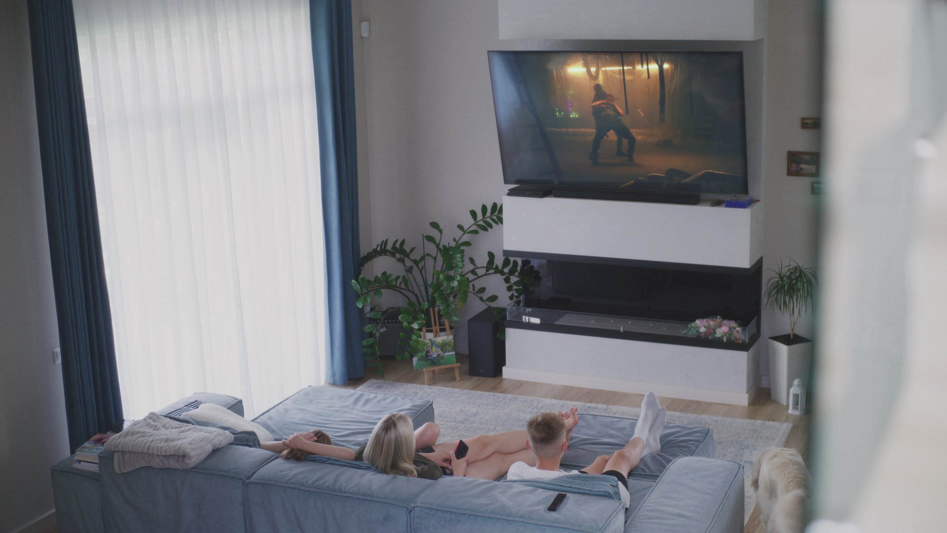 Mother with her children rest on couch in light living room and watch action movie, dog comes into room and sits near. Big TV screen with displayed film. Family spend weekend together. High angle.