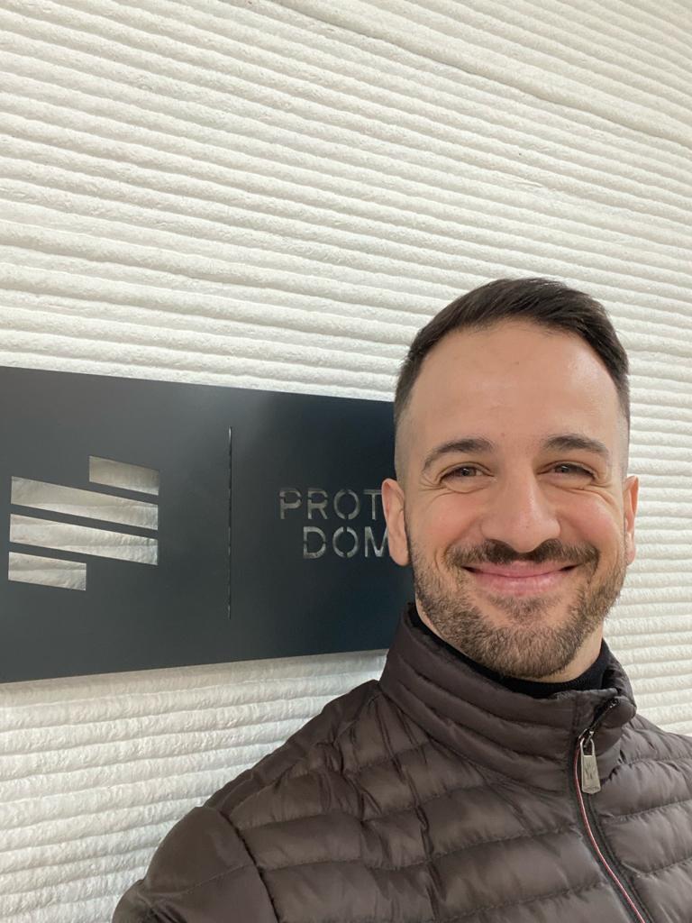 Person smiling in front of a sign with the logo and partially visible text, wearing a quilted jacket against a textured wall.