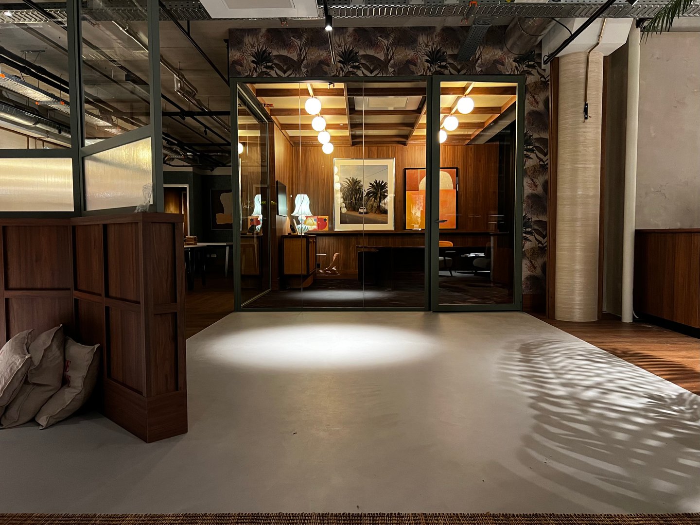 Modern office space with glass walls, pendant lights, and tropical-themed decor. Wooden floors and a stylish, cozy ambiance.