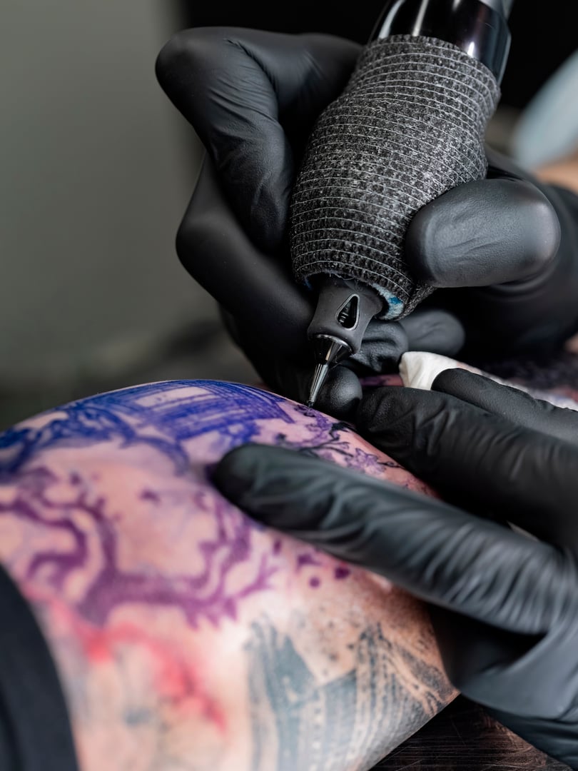 Extreme close-up of tattoo machine grip and needle cartridge positioned over colorful Japanese landscape design with purple stencil.