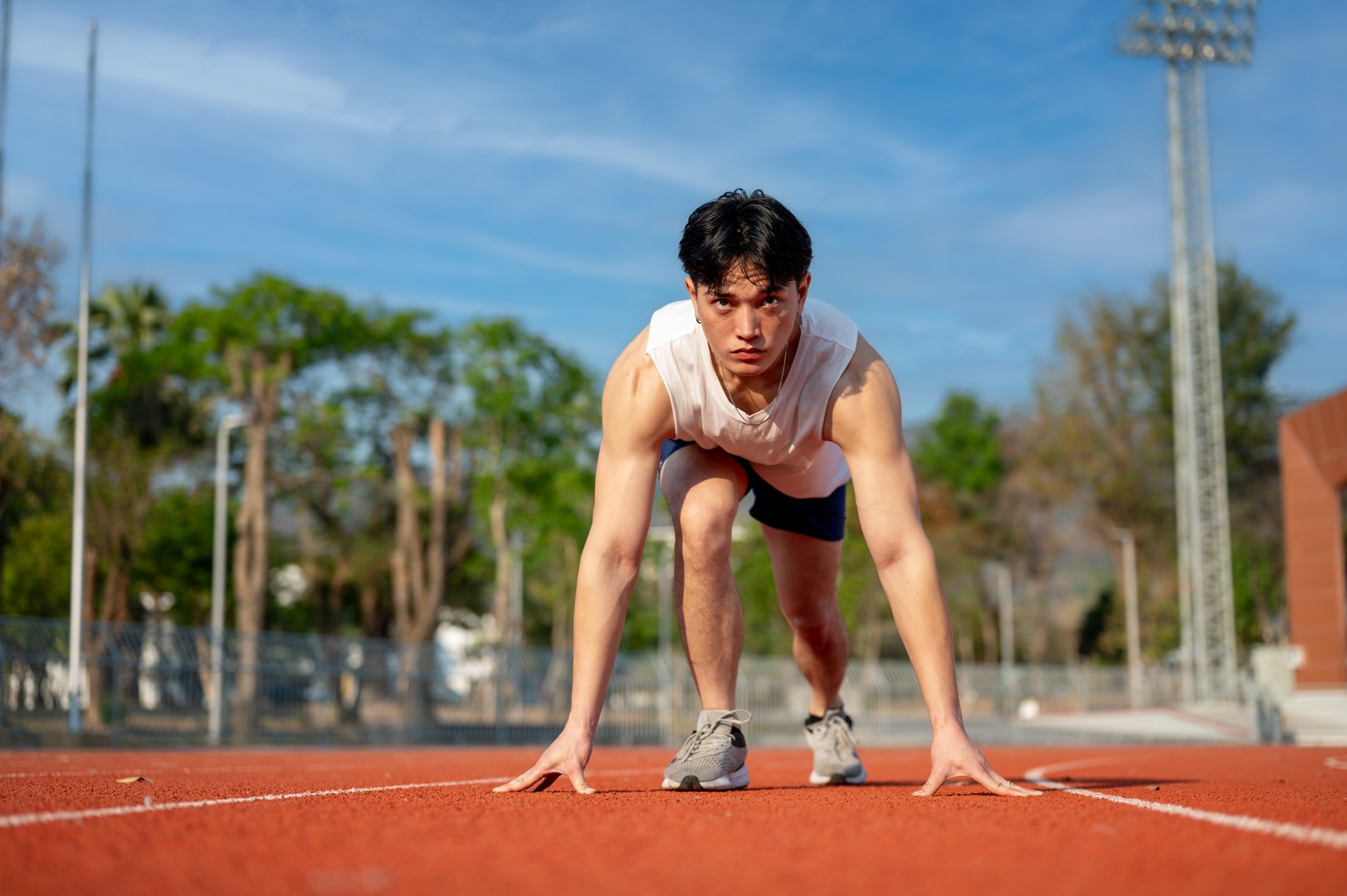 Asian man in white tank top is crouching in a set position ready to run on racetrack. Outdoor exercise or work out, Athlete training, Sport and health.