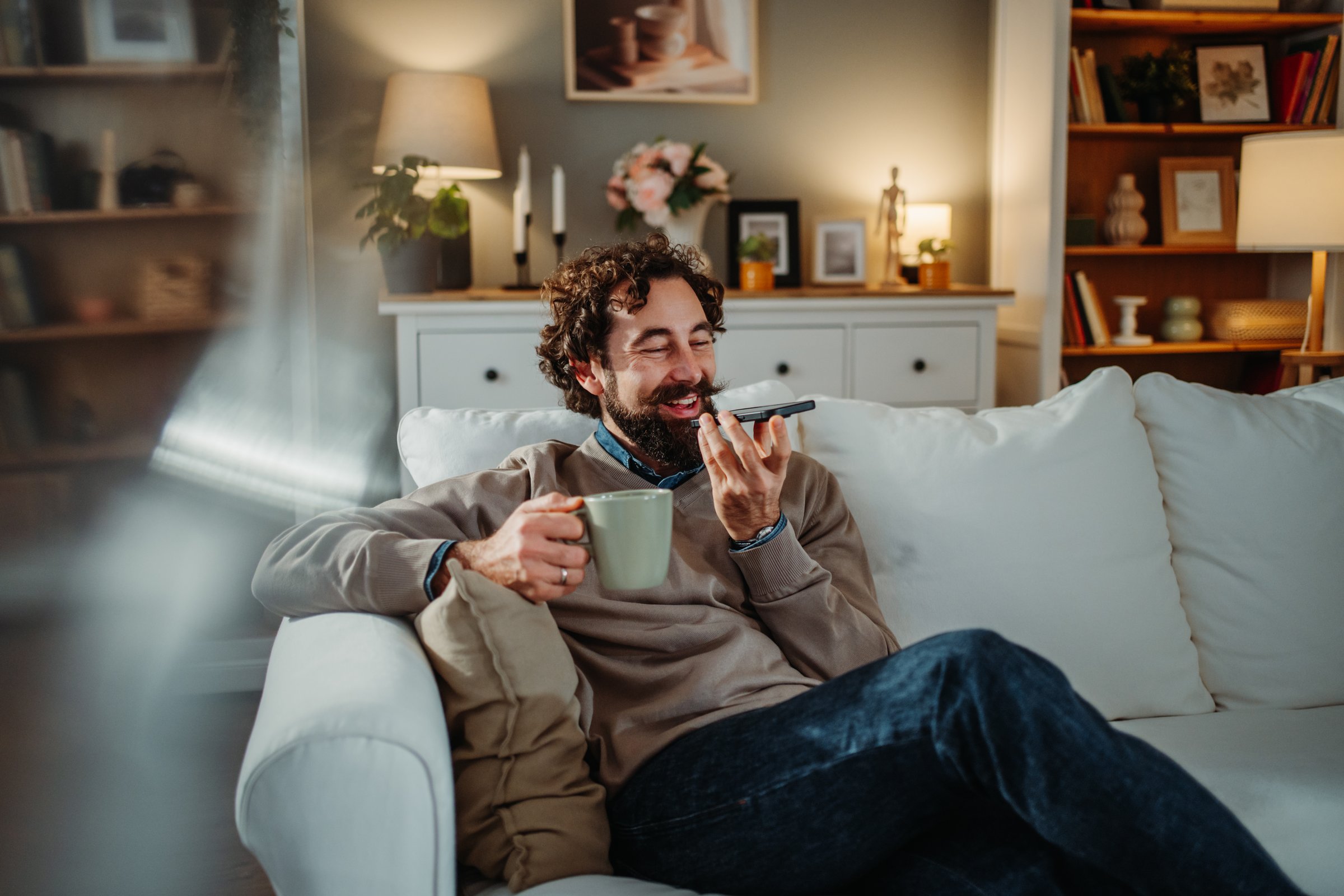 Smiling man relaxing on sofa while dictating voice message on smartphone at home