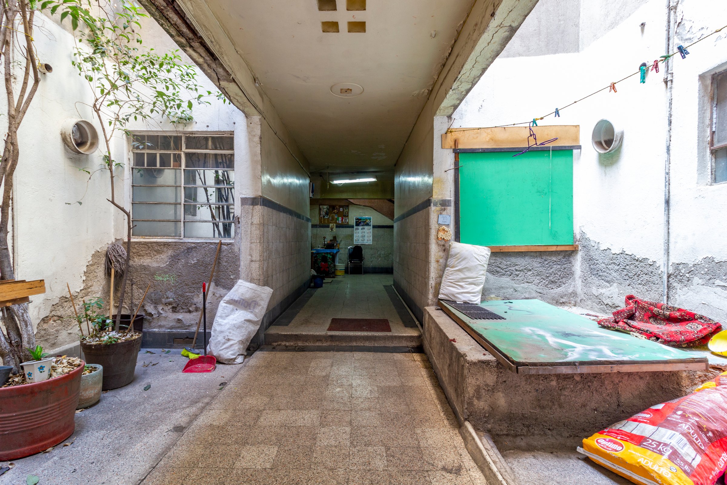 A narrow, covered passageway with a potted plant, cleaning supplies, and bags, leading to a small, cluttered interior space.