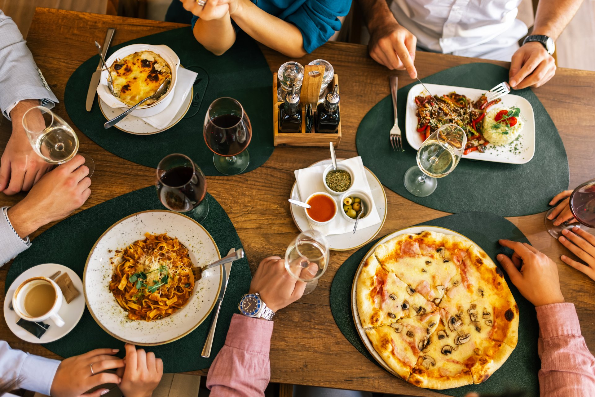 Top view of hands at a restaurant lunch, with pizza, pasta, rice, chicken, vegetables, and wine on the table