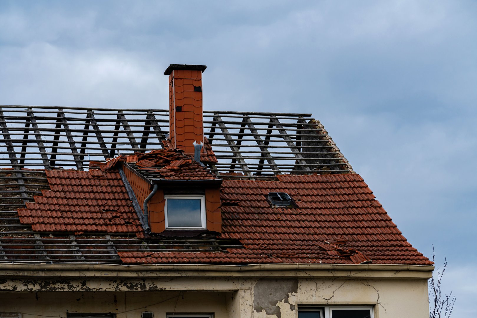 The roof shows significant damage with broken tiles and an exposed structure. The chimney remains intact, while dark clouds loom overhead, hinting at possible further inclement weather.