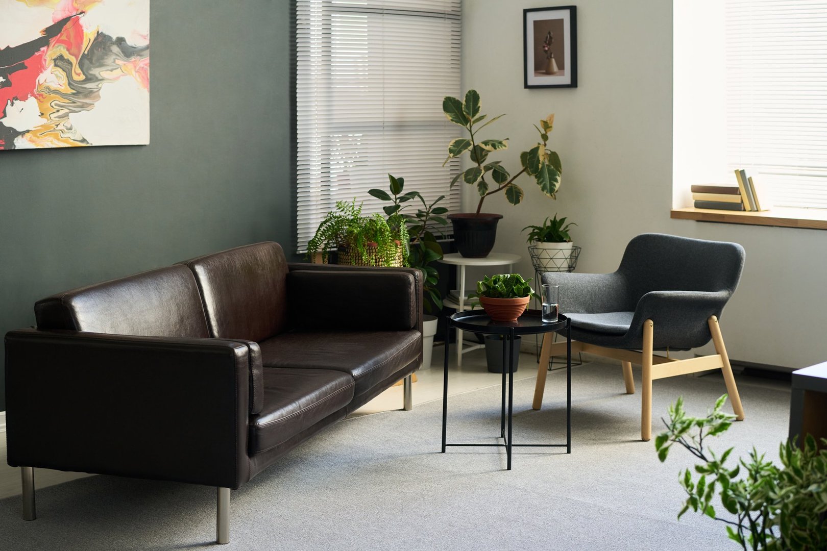 Empty psychologist office showing modern waiting area with leather sofa, armchair, potted plants and abstract artwork on wall, sunlight streaming through window blinds