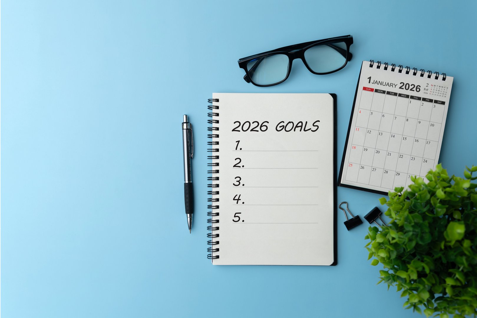 Notebook titled 2026 Goals with a blank checklist on a blue desk beside pen, glasses, mini calendar, representing life and business goal planning, strategy, productivity and a fresh start.target