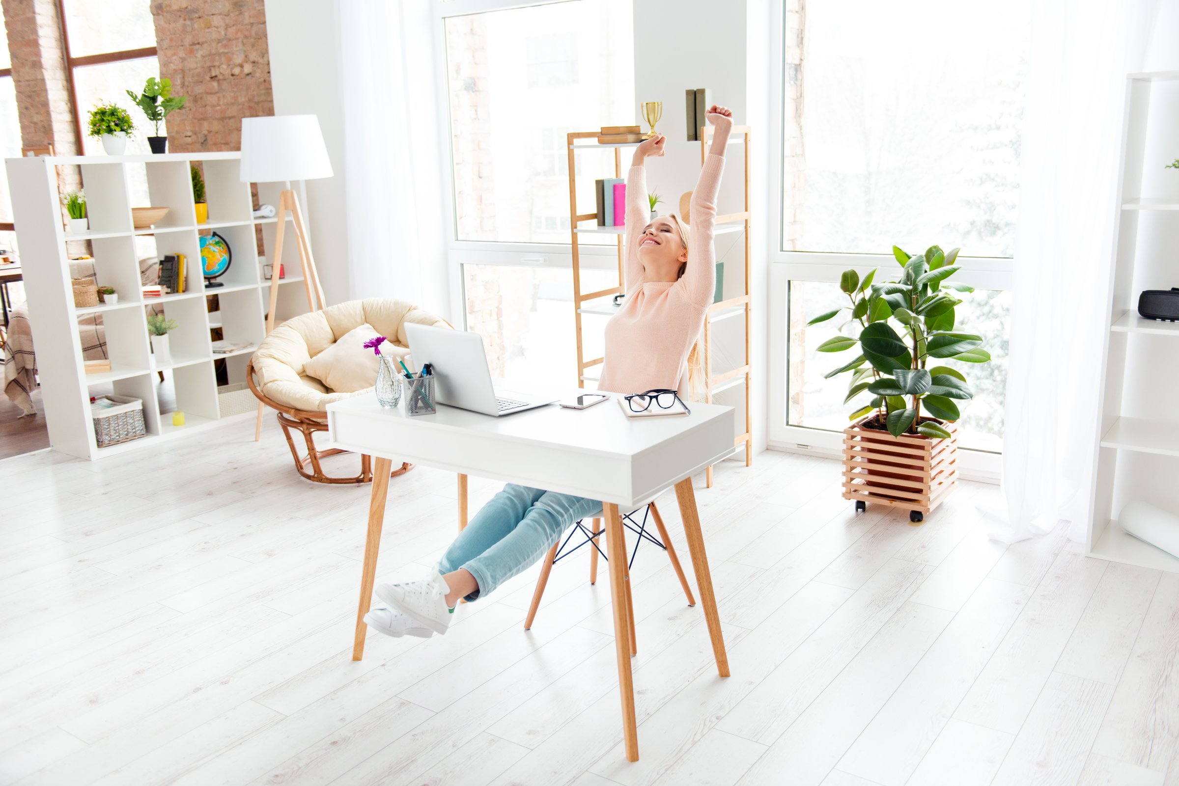 Portrait of cheerful positive girl sitting in white room at desktop making stretching with raised arms while preparing for exams enjoying timeout dreaming about weekend holiday