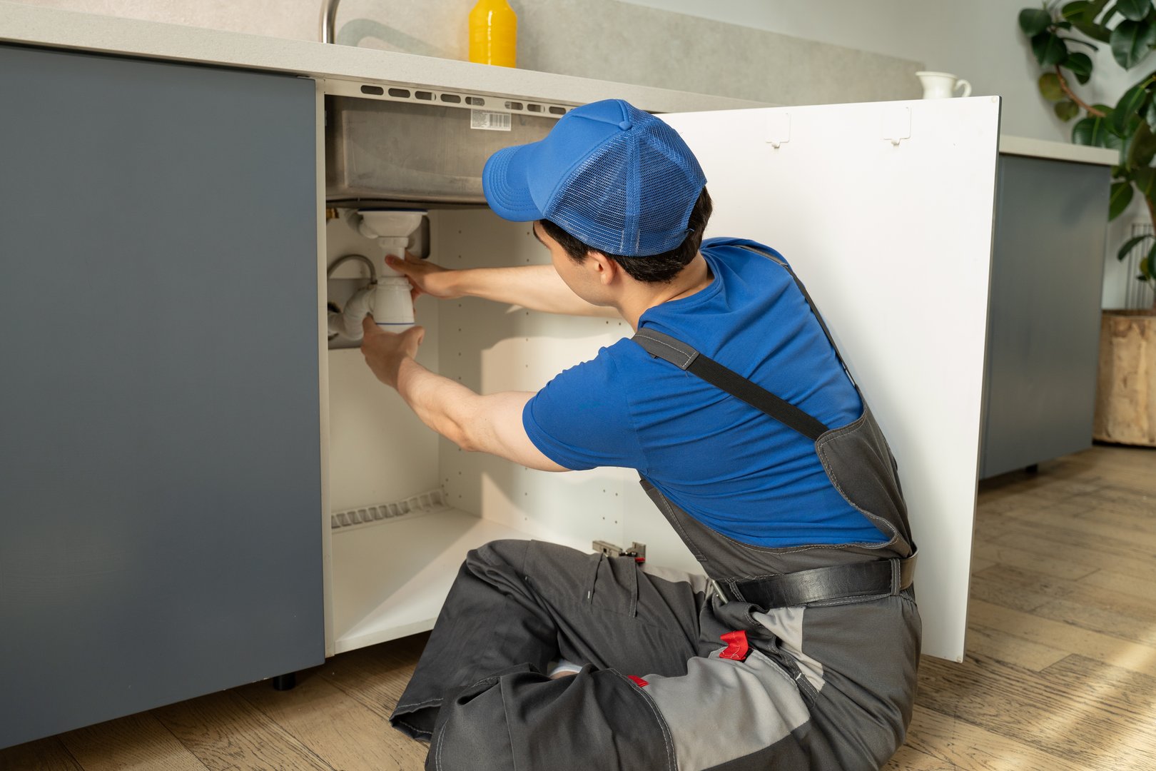 A skilled plumber in a blue uniform and cap is working diligently to repair the pipes located under the sink in a well-lit, contemporary kitchen setting, equipped with various tools for the job.