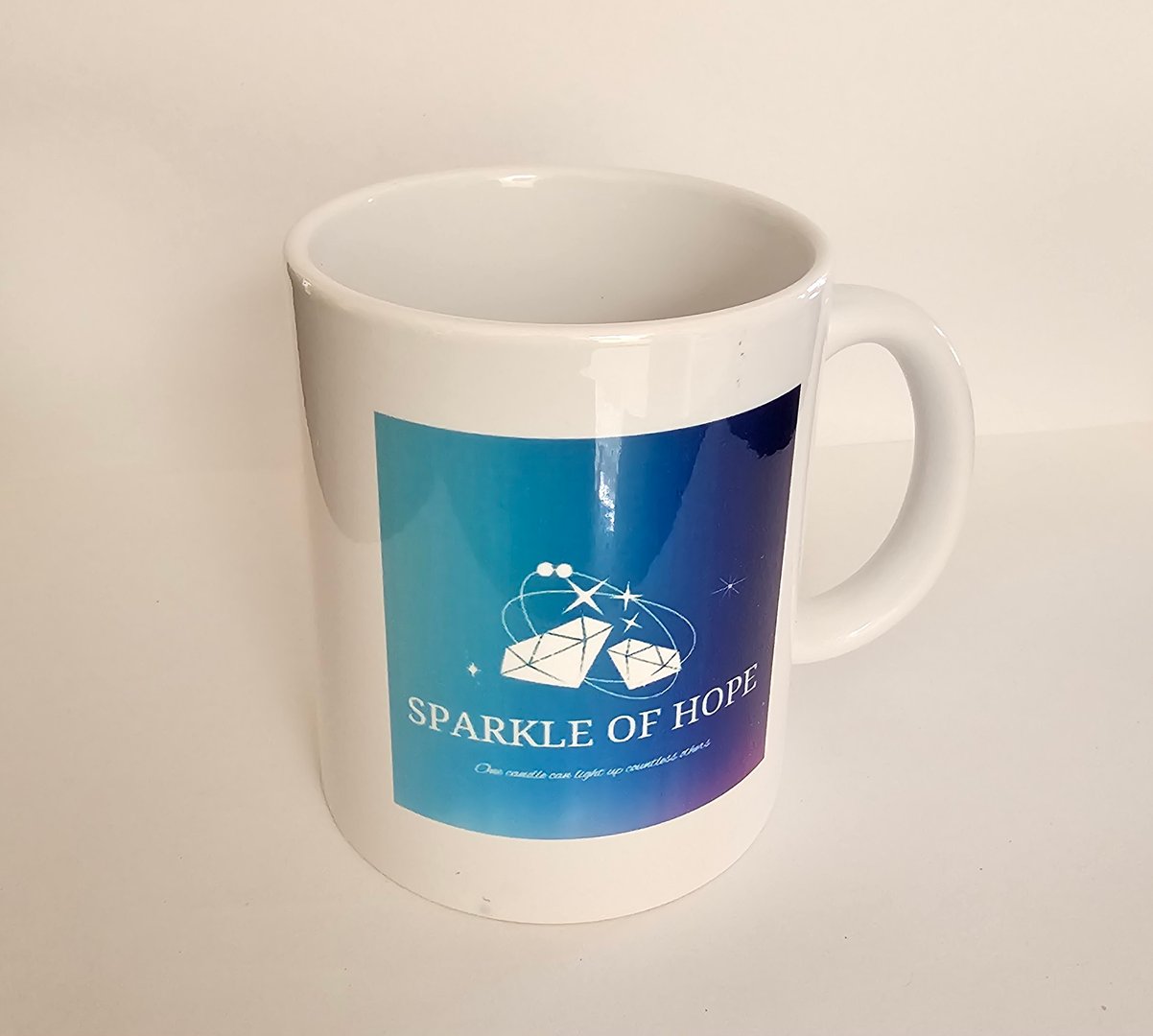 White mug with 'Sparkle of Hope' logo featuring geometric design on a blue gradient background.