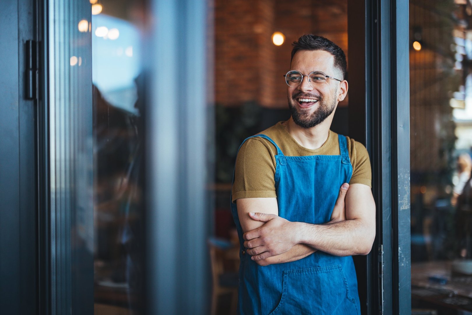 Smiling male bartender, clad in apron, stands arms crossed in a cozy coffee house, ready to serve a diverse clientele with a variety of coffee beverages.