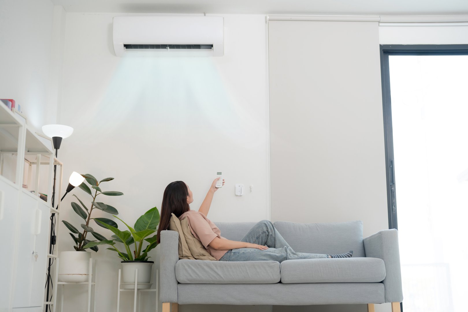 Content young woman lounges on a sofa, holding a remote to adjust her wall-mounted air conditioner, appreciating the cool air in a modern, bright living room setting