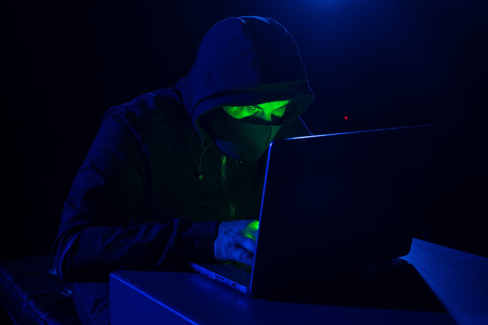 Cyber hacker in hood behind laptop in dark room, digital cyber security concept, computer hacking, information security